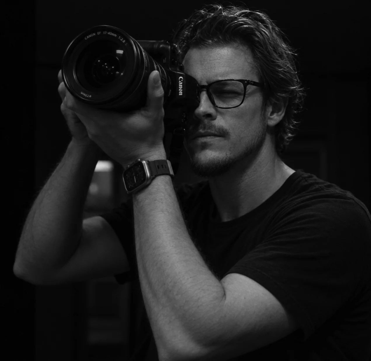 Bret Lindquist holding a camera to his eye in a black and white portrait.
